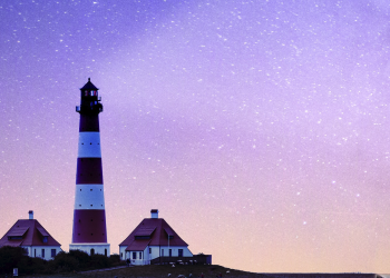 A lighthouse rises above two houses at twilight. The sky is full of stars, and transitions smoothly from dark blue at the top, to light purple in the middle, to orange-pink near the horizon. The lighthouse, is a narrow cone, slightly thicker at the bottom than the top. It's painted in wide horizontal stripes of red and white, and it casts a beam of light across the sky.